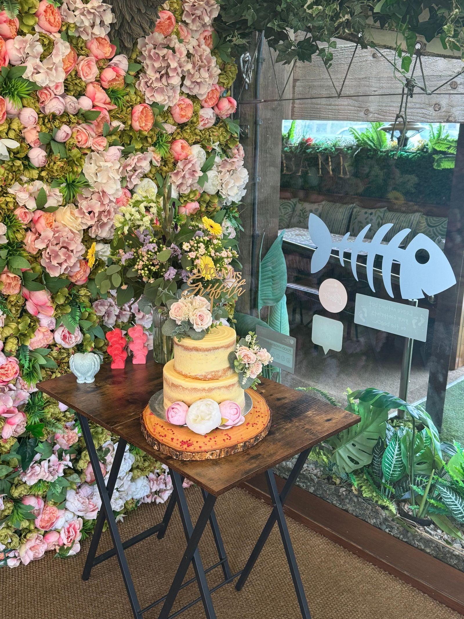 A semi-naked celebration cake topped with fresh flowers against the floral wall at The Bank at Bude