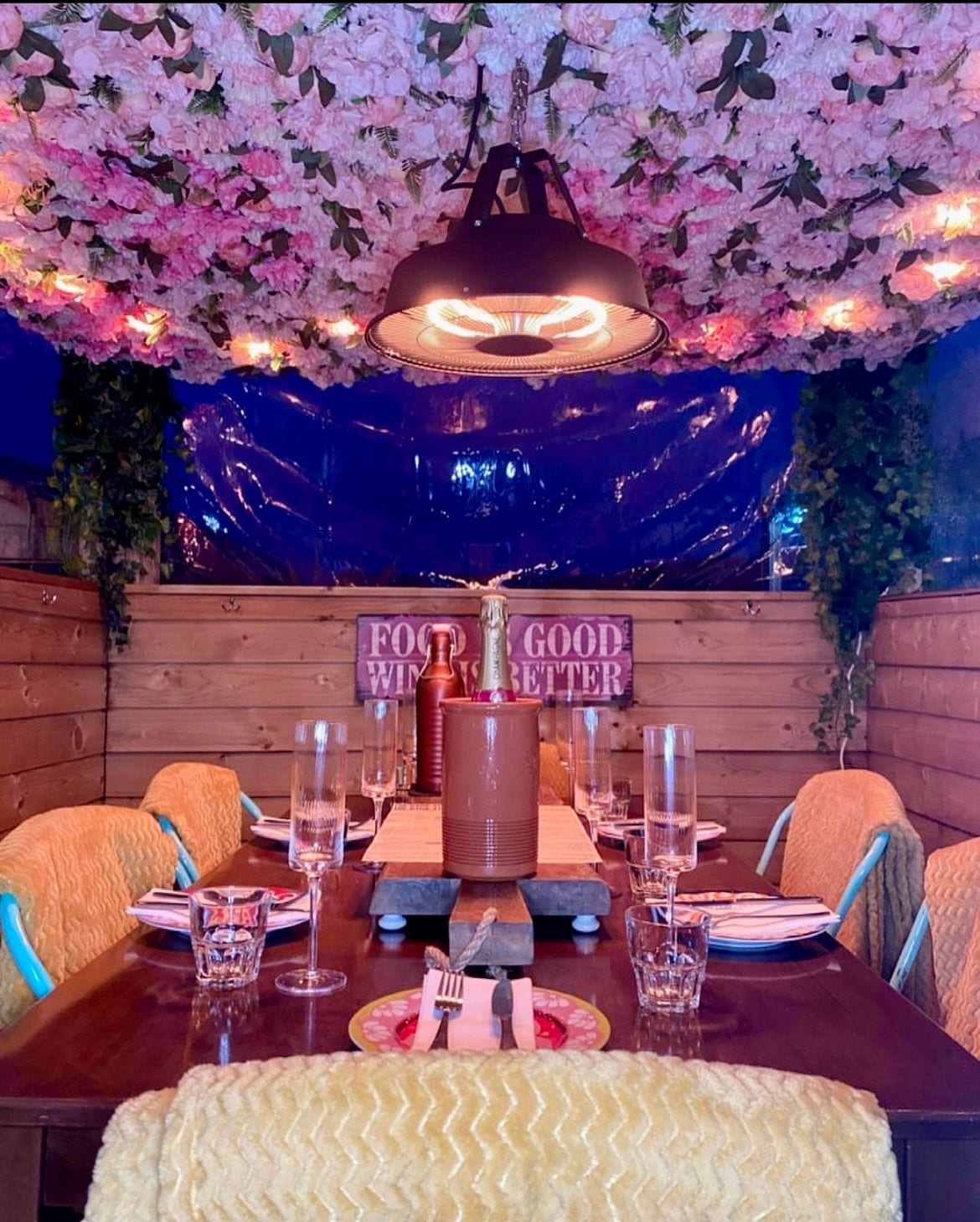 A riverside dining pod at The Bank at Bude set for a group celebration with Champagne, glasses and cosy blankets along a long table beneath the lit flower-canopy ceiling