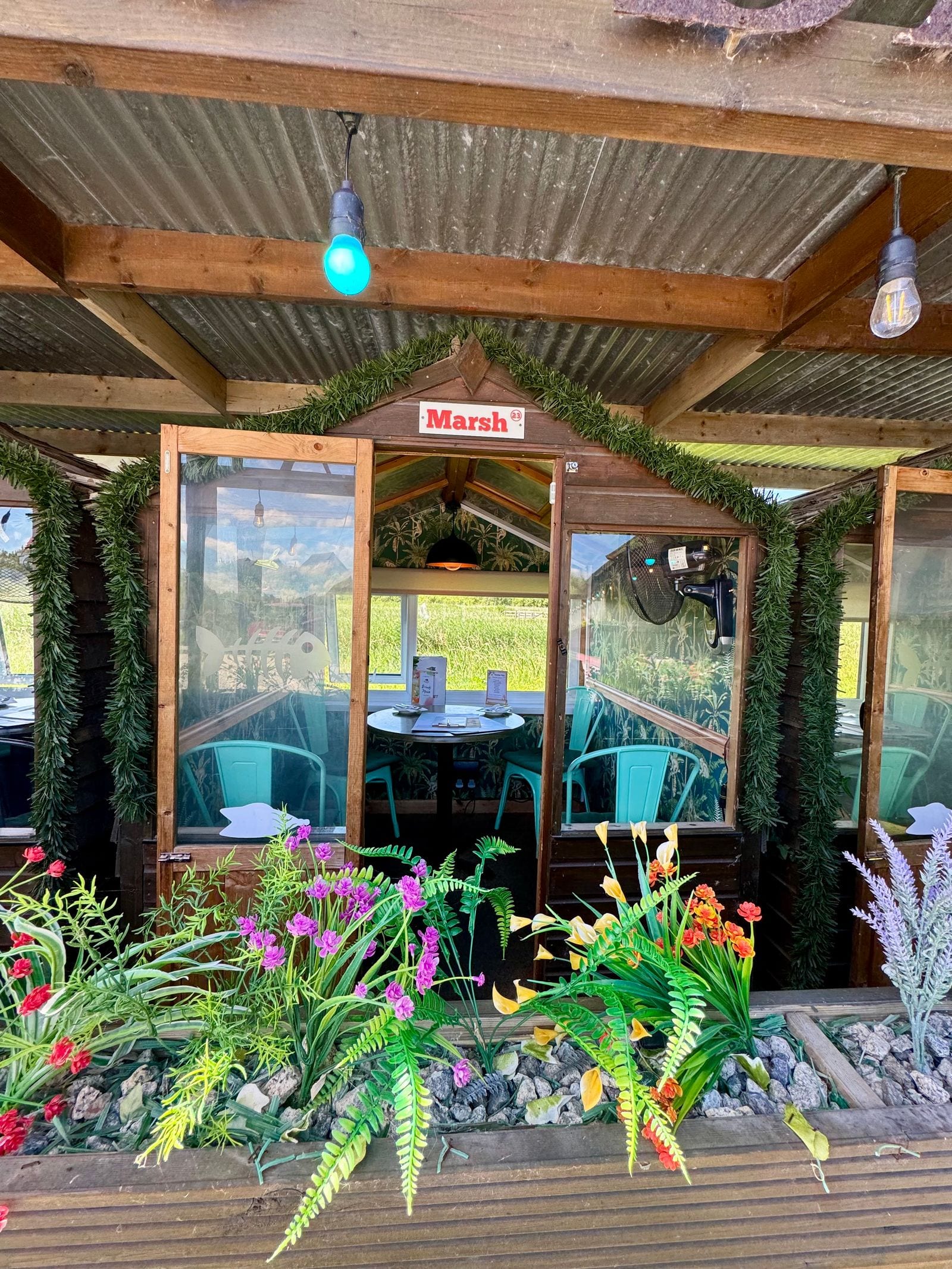 The ‘Marsh’ riverside dining pod at The Bank at Bude in summer, framed by bright flowers, with the River Neet meadows beyond