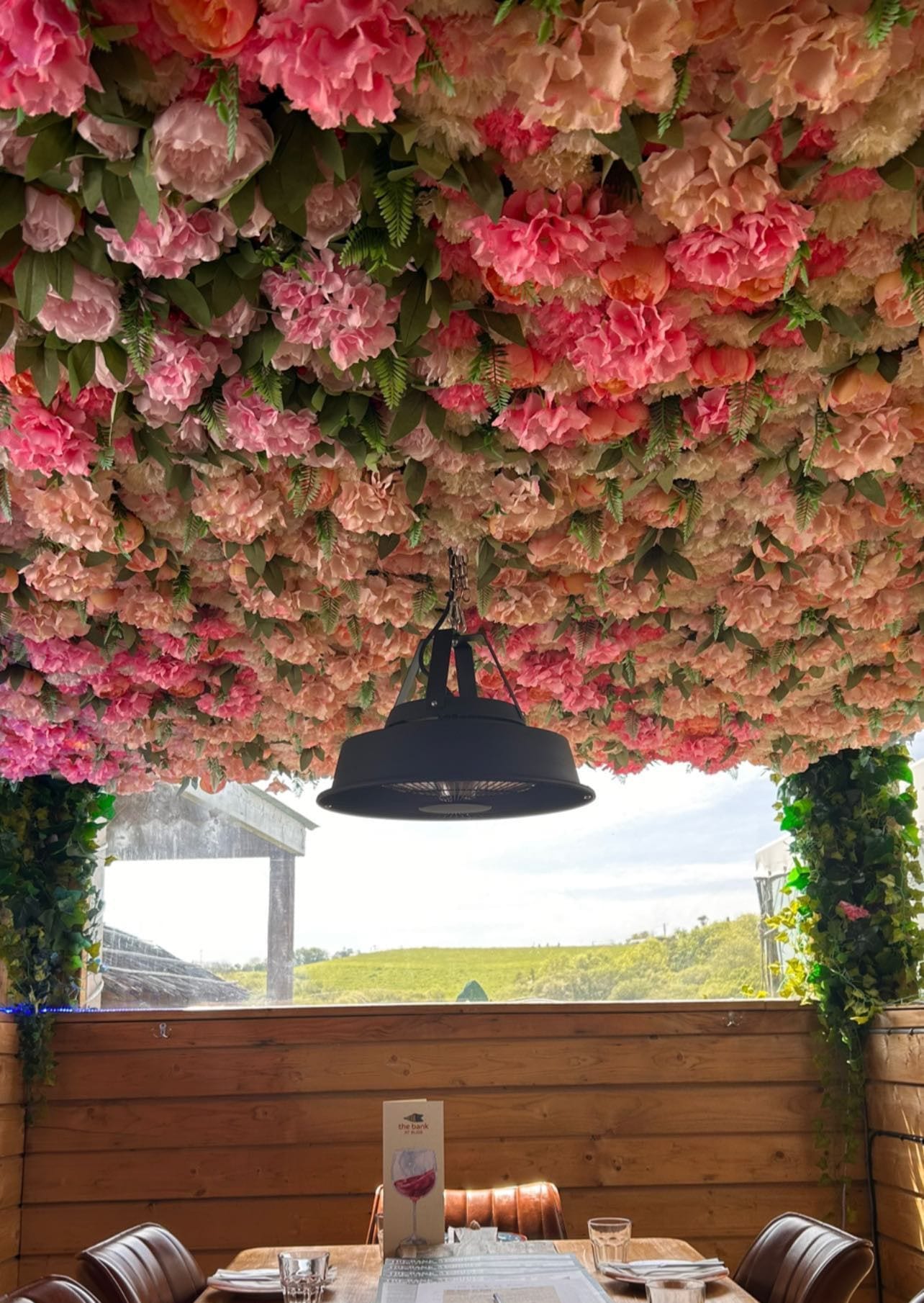 The XL riverside dining pod at The Bank at Bude beneath its pink flower-canopy ceiling and hanging heater, with countryside views over the River Neet marsh
