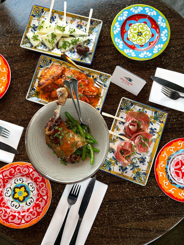 Overhead tapas spread on a wooden table with hand-painted Spanish ceramic plates and a The Bank at Bude branded napkin