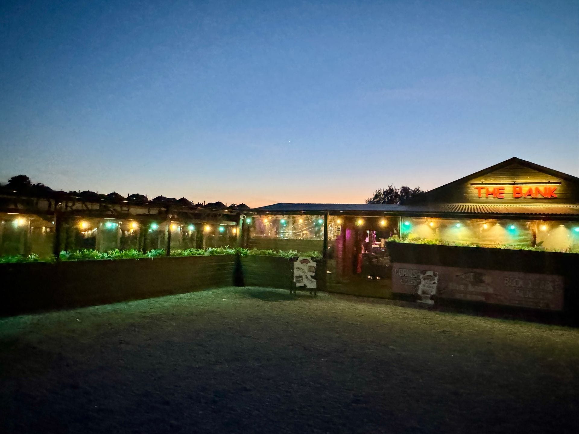 The Bank at Bude lit at dusk beside the River Neet, with the terrace dining pods glowing under a sunset sky
